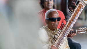 Baluji Shrivastav OBE smiling and holding the sitar. He is wearing gold and is wearing dark glasses. 