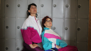 Candoco - Kimberley and Anne-Gaelle are in front of a block of silver school lockers. They are both wearing early 90s shell suits. Kimberleys shell suit is turquoise, fuchsia and white, and Anne-Gaelles shell suit is Fuchsia and white with a blue, pink, and white detailing in a downward v-shape. Both Kimberley and Anne-Gaelle have their hands on their hips, looking at the camera with a serious expression. 