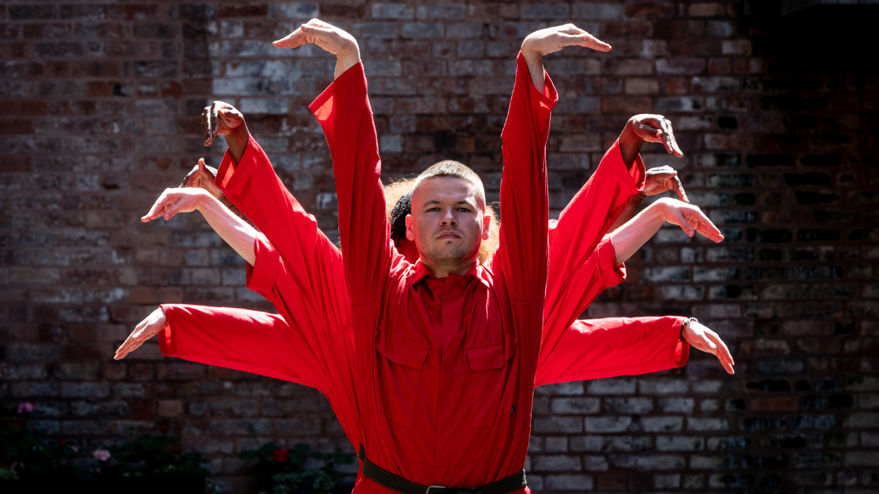 Billy Read is facing the camera in a red boiler suit with his arms above his head and hands facing out. There are 4 people stood behind him, with their arms and hands out at different angles