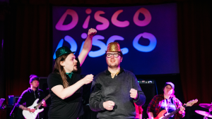 Two performers are on stage, they both wear sparkly hats and bow ties, one green, one gold. One appears to be showing the other and imaginary scroll. Behind them musicians play and a giant screen says DISCO DISCO in pink and purple letters.
