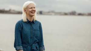 Ellen (woman in her twenties with albinism) standing by the sea, wearing a blue jumpsuit, laughing at the camera