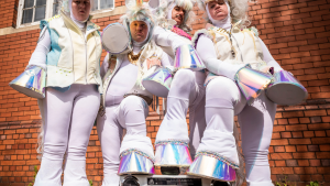 Four people dressed as unicorns standing on two legs. They look grumpy and have one of their hooves raised on a boombox