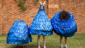 Three Janes are in a line in a garden with a red brick wall behind them. One Jane is crouched down underneath their big blue skirt so it looks like they are a walking skirt. Beside her is another Jane who is standing upright with their fingertips all jagged shooting up into the air with the skirt tied around the wrists, covering the face. The third Jane has the skirt around her face, so all you see is hair peeping through where it should be. 