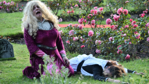 A young man, wearing a pink dress and blonde wig is kneeling next to a woman who is lying on the ground. In the background is a small tombstone and the man in the dress is crying in a funny way.