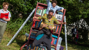 Four performers wearing brightly coloured shorts and glasses. One performer stands at the side, holding a book. One performer is holding two other performers. They are gripping on with their arms and legs and have created a creature with their bodies! Their hands have made claws and their faces are snarling.  A filled bookshelf is in the background.
