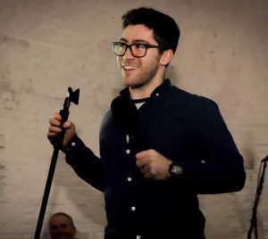 Aaron Simmonds - A man with black glasses and a black shirt smiles as he holds onto a microphone stand.