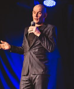 Steve Day - A man wearing a smart black suit stands on stage speaking into a microphone. The stage is bathes in blue light