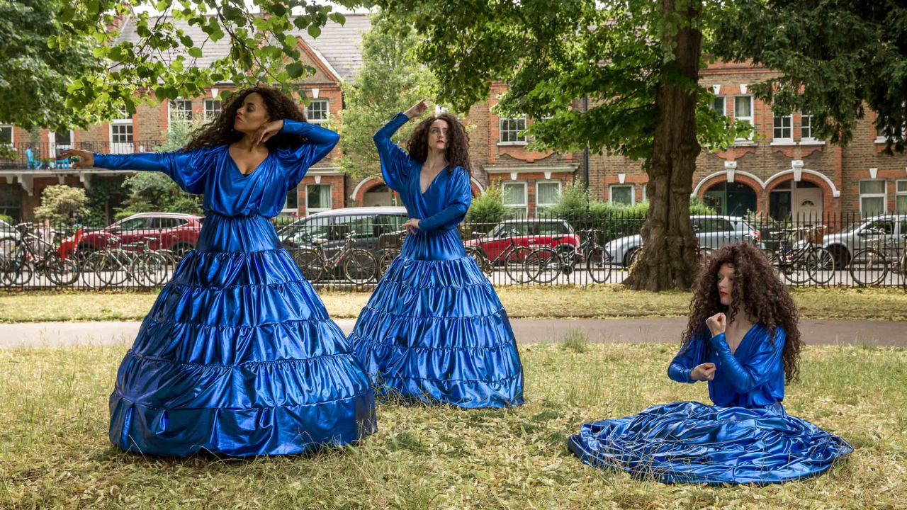 3 dancers with big curly brown wigs and metallic blue hoop skirts are in a park. They are doing poses taken from pre Raphaelite paintings.