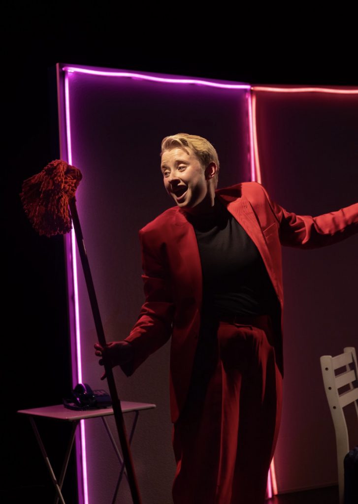 Hannah Maxwell on stage in a red suit with black top holding a mop with a telephone table and chair in the background. 